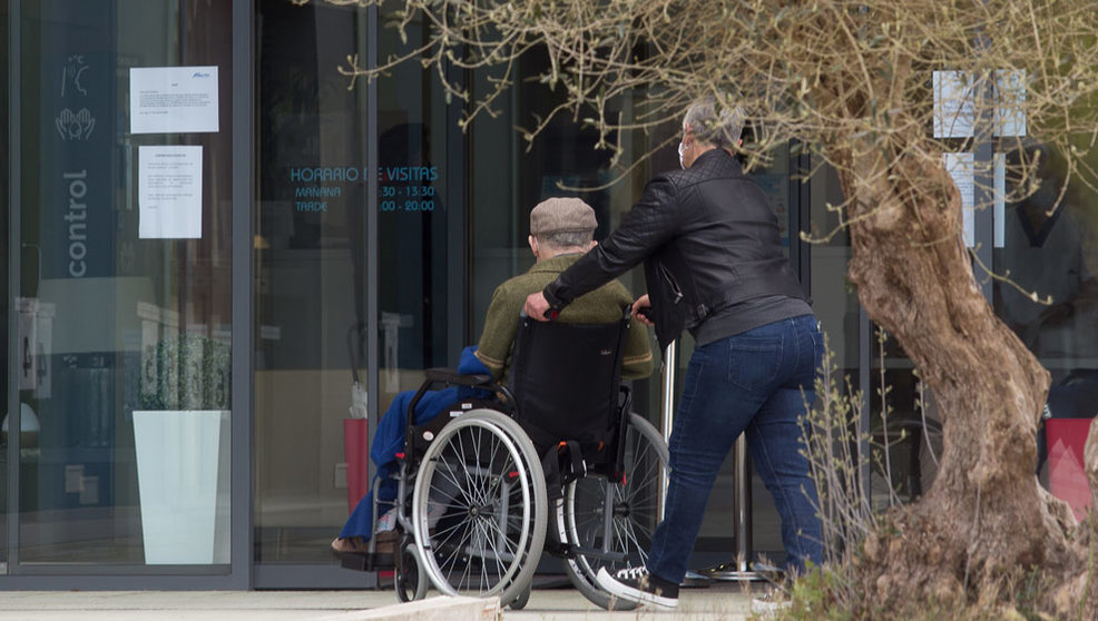 Una mujer pasea junto a un anciano en silla de ruedas hacia una residencia