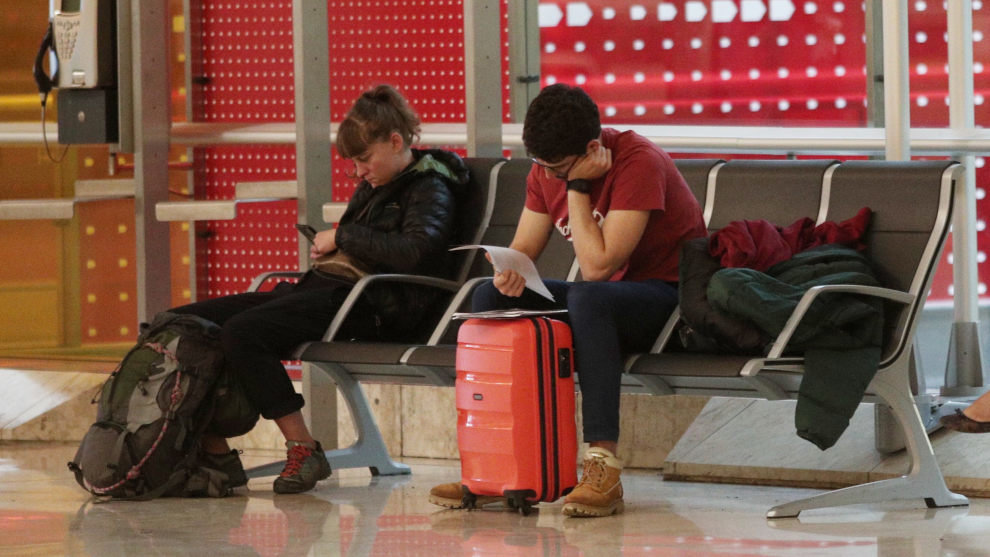 Dos personas con su equipaje esperan su vuelo sentados en un banco en el Aeropuerto de Madrid-Barajas Adolfo Suárez